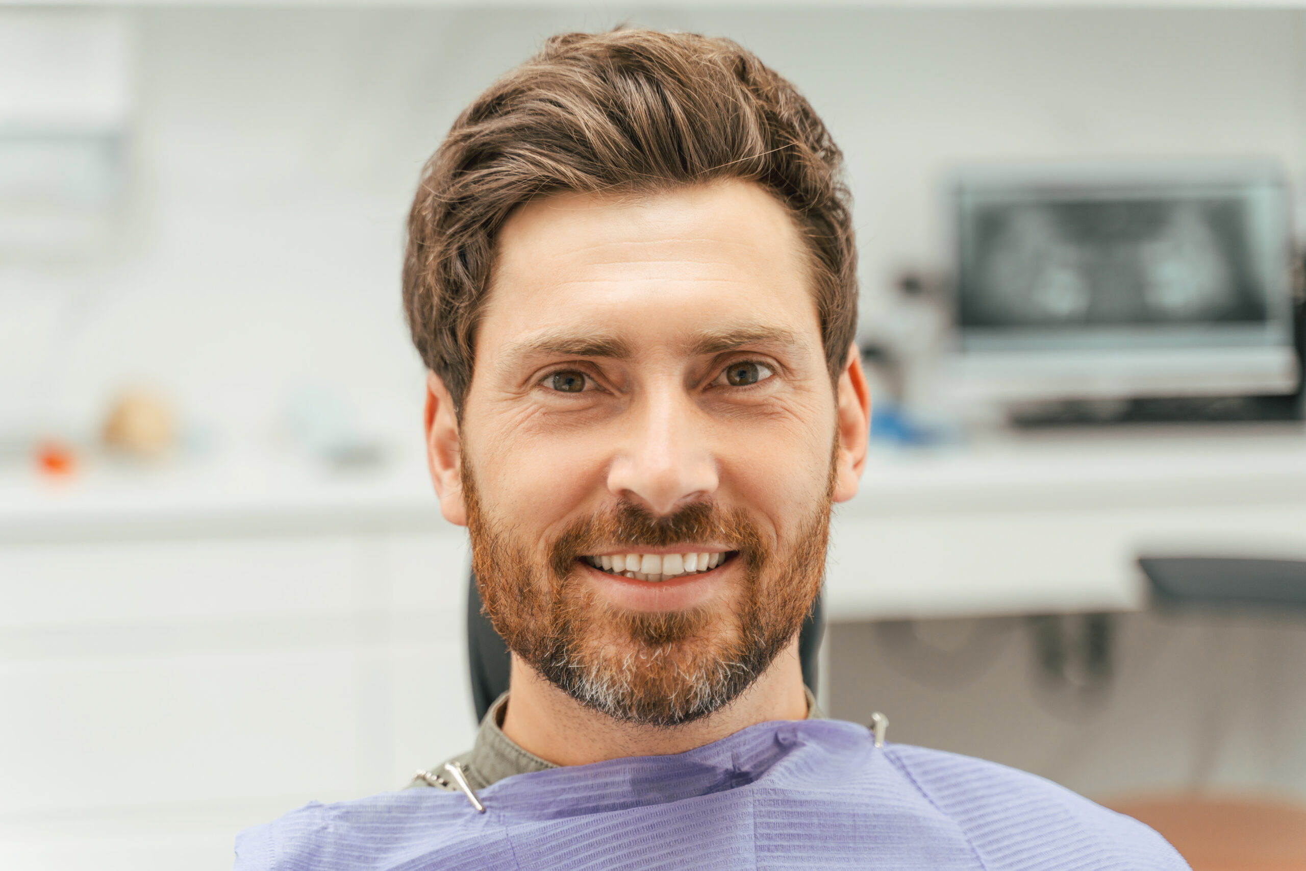 Attractive smiling bearded man sitting in dental chair, looking at camera.