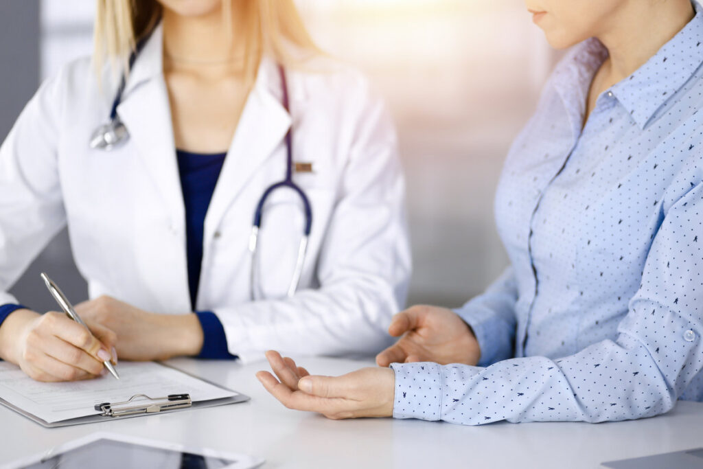 Unknown woman- doctor is listening to her patient, while sitting together at the desk in the sunny cabinet in a clinic. Female physician with a stethoscope is writing at clipboard, close up. Perfect medical service in a hospital. Medicine concept.